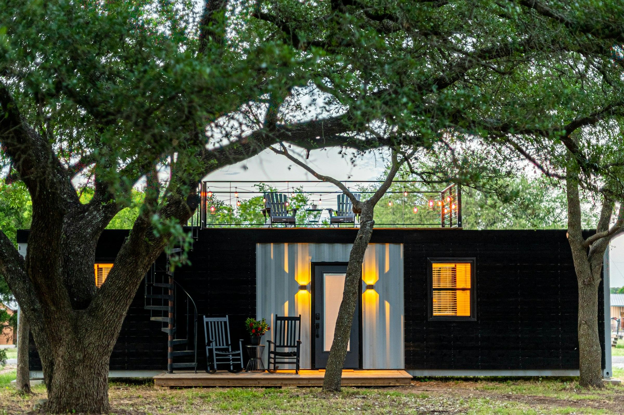 A black container home glowing warmly at dusk beneath old trees
