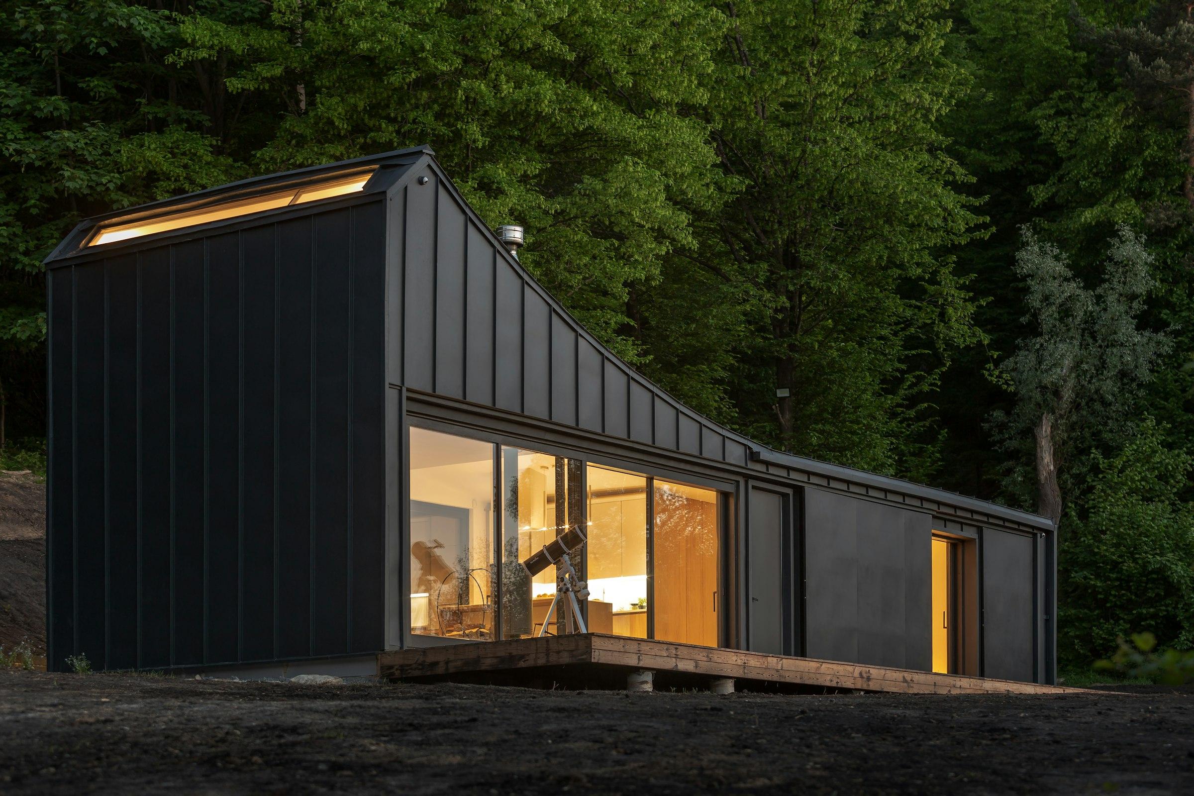 A black-clad modular cabin glowing at dusk in a forest clearing